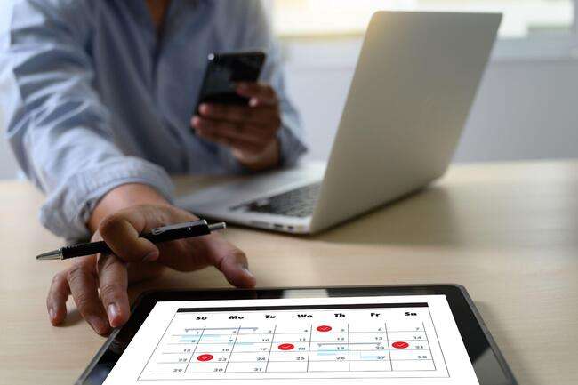 Person using a tablet to check a calendar with red dots on selected dates, holding a pen in one hand and a phone in the other, with a laptop in the background.