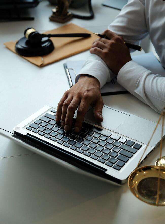 A lawyer's hand on a laptop keyboard with legal documents and a gavel on the desk.