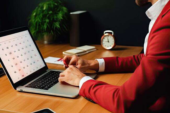 A professional wearing a red blazer works on a laptop, displaying a digital calendar on the screen. An analog clock is visible in the background, signifying time management and scheduling.