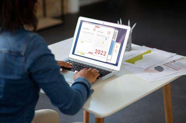 A person in a blue denim shirt is working on a laptop, displaying a calendar for the year 2022. Various documents and a pen are visible on the desk, indicating a well-organized workspace.