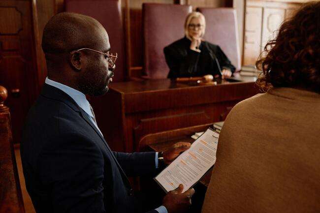 A lawyer in a courtroom, attentively reviewing documents, while a judge is visible in the background, focusing on the proceedings.