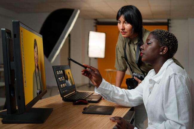 Two women collaborating on a project using a desktop computer and laptop, representing teamwork in immigration case management.