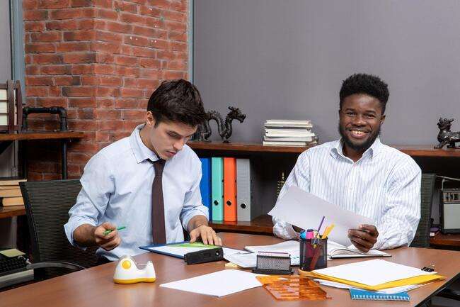Two young professionals working at a desk with various office supplies and documents.